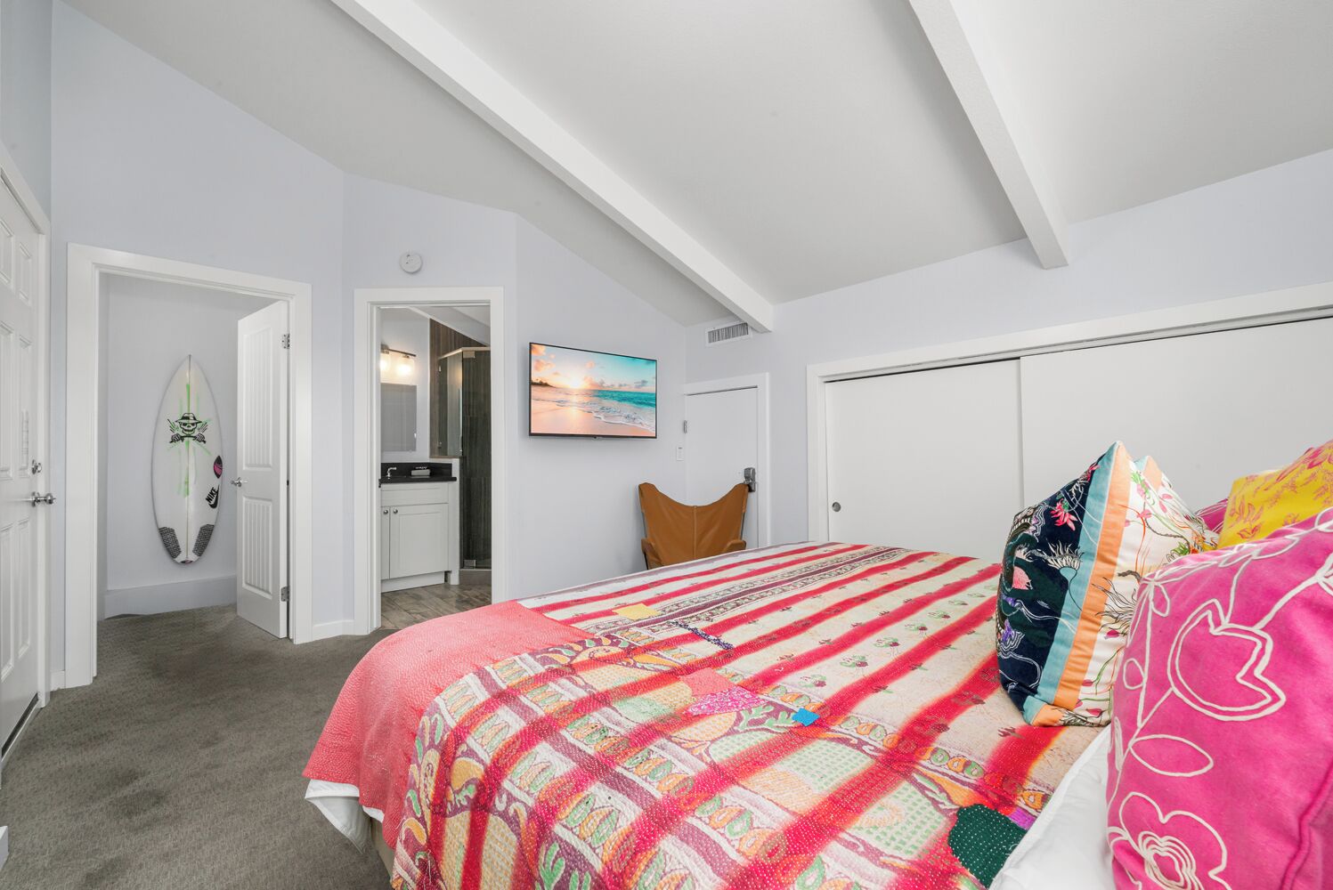 A bedroom with a bed, a colorful quilt, a surfboard, a painting, and a closet.
