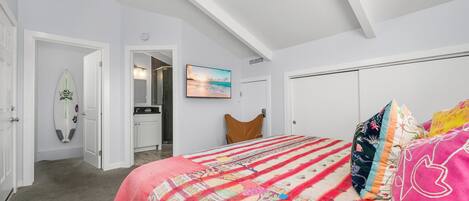 A bedroom with a bed, a colorful quilt, a surfboard, a painting, and a closet.