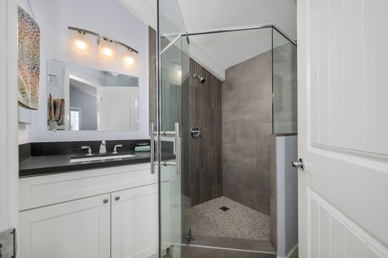 Une salle de bain moderne avec une douche à l’italienne en verre, un meuble-lavabo blanc doté d’un plan de travail noir et un grand miroir au-dessus du lavabo.
