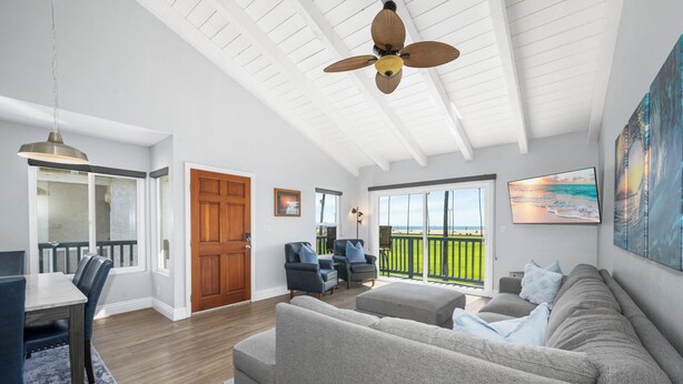 A spacious living room with a large gray sofa, a dining area with a table and chairs, a ceiling fan, and a view of the beach through a sliding glass door. - 2 Beachfront Condos with Private Balcony (Newport Beach)