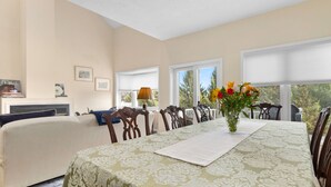 Une salle à manger avec une table dressée pour six personnes, entourée de chaises en bois. Un vase rempli de fleurs est posé sur la table, et la pièce est dotée de grandes fenêtres équipées de stores.