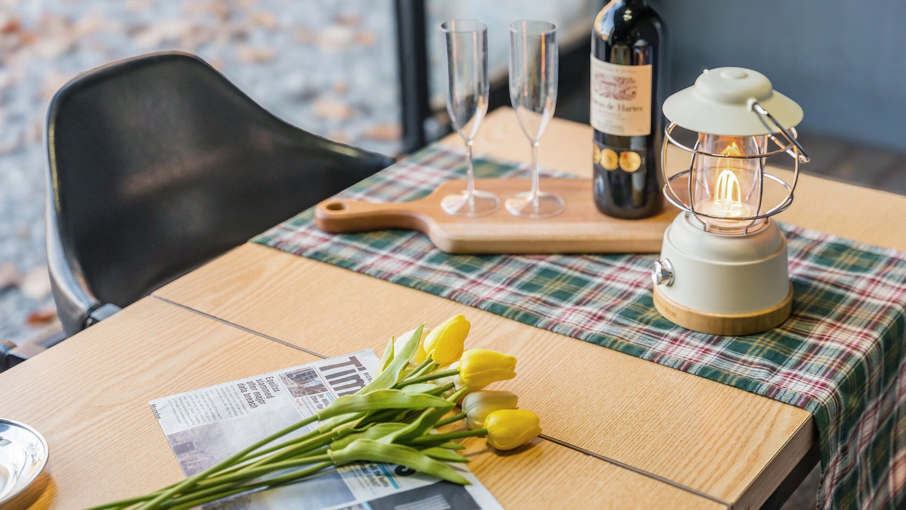 Una mesa con un periódico, un ramo de tulipanes amarillos, una botella de vino, dos copas y un farol.