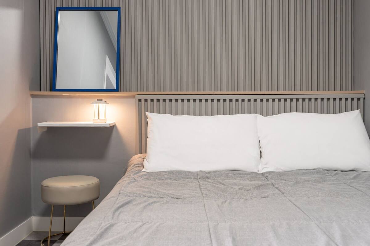 Un dormitorio ordenado con cama, una mesita de noche con lámpara, un pequeño taburete y un estante en la pared.