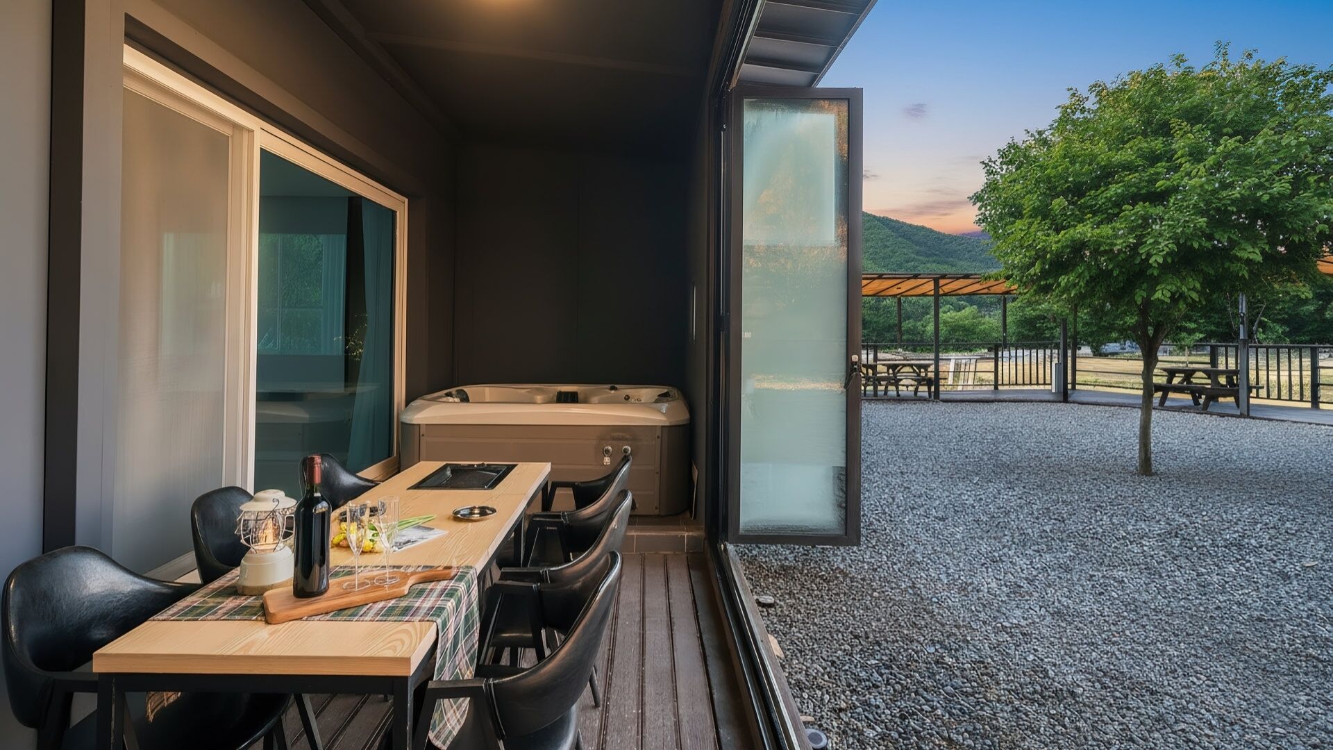 Un balcón con jacuzzi, mesa de comedor y sillas, con vistas a un área de grava con árboles y una vista distante.