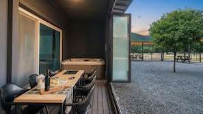 Un balcón con jacuzzi, mesa de comedor y sillas, con vistas a un área de grava con árboles y una vista distante.