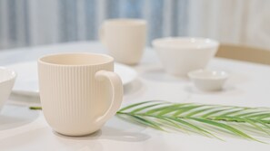 Une table blanche sur laquelle se trouvent plusieurs tasses et bols en céramique blanche, une assiette blanche et une feuille de palmier verte.