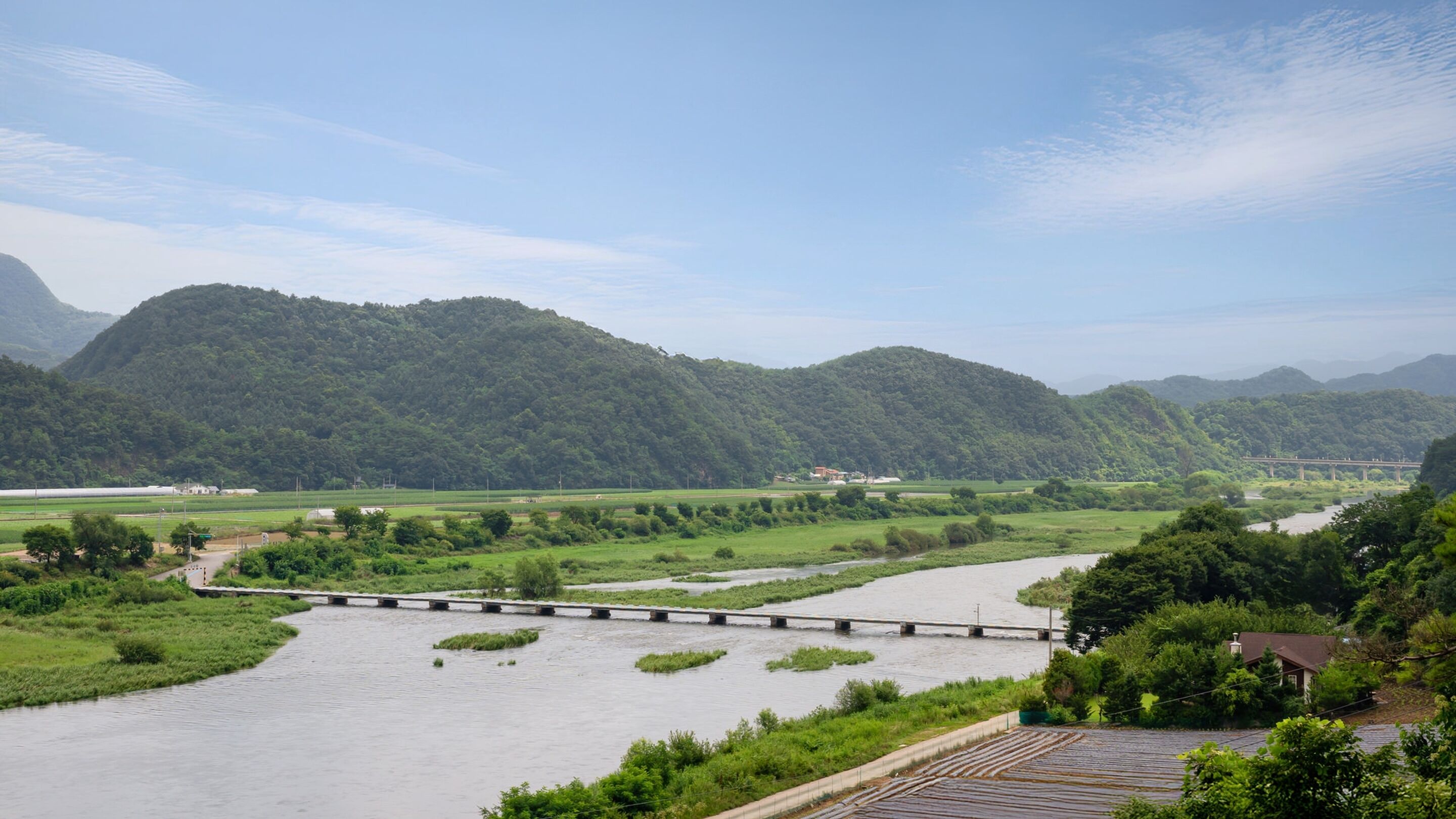 A river with a bridge, lush green hills, and a few buildings in the distance.