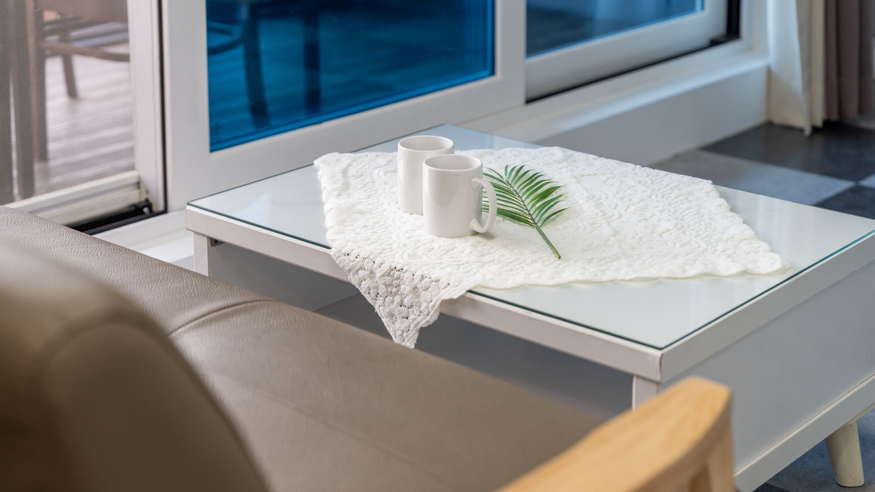 Ein modernes Wohnzimmer mit einem Glastisch, weißen Kissen und Blick ins Freie durch große Fenster.