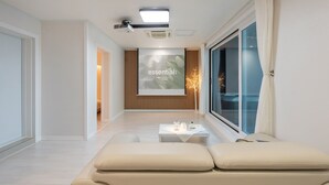 A modern hotel room with a large window, a cozy seating area with cushions, a small table with amenities, and a wall-mounted projector screen displaying 'essential;'. - Yeongwol You and I Spa Pension (Yeongwol)