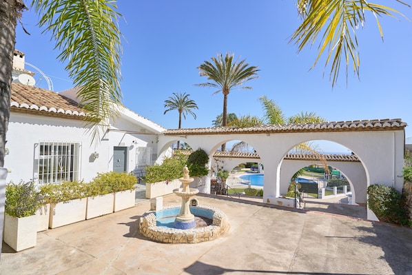 A courtyard with a fountain, palm trees, and a pool visible through an archway.