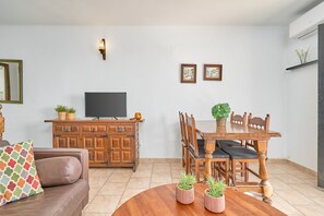 A living room with a sofa, TV, wooden cabinet, dining table, chairs, and potted plants.
