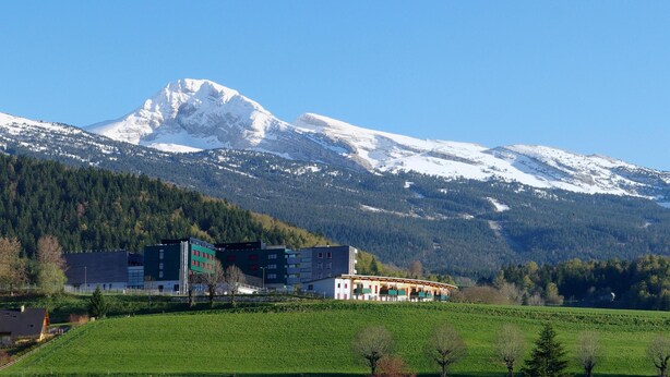 Een berg met besneeuwde toppen, een groen veld en een gebouw met meerdere verdiepingen en balkons.