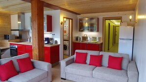 Een woonkamer met een bank en een salontafel, een keuken met rode kastjes, en een wasmachine.
