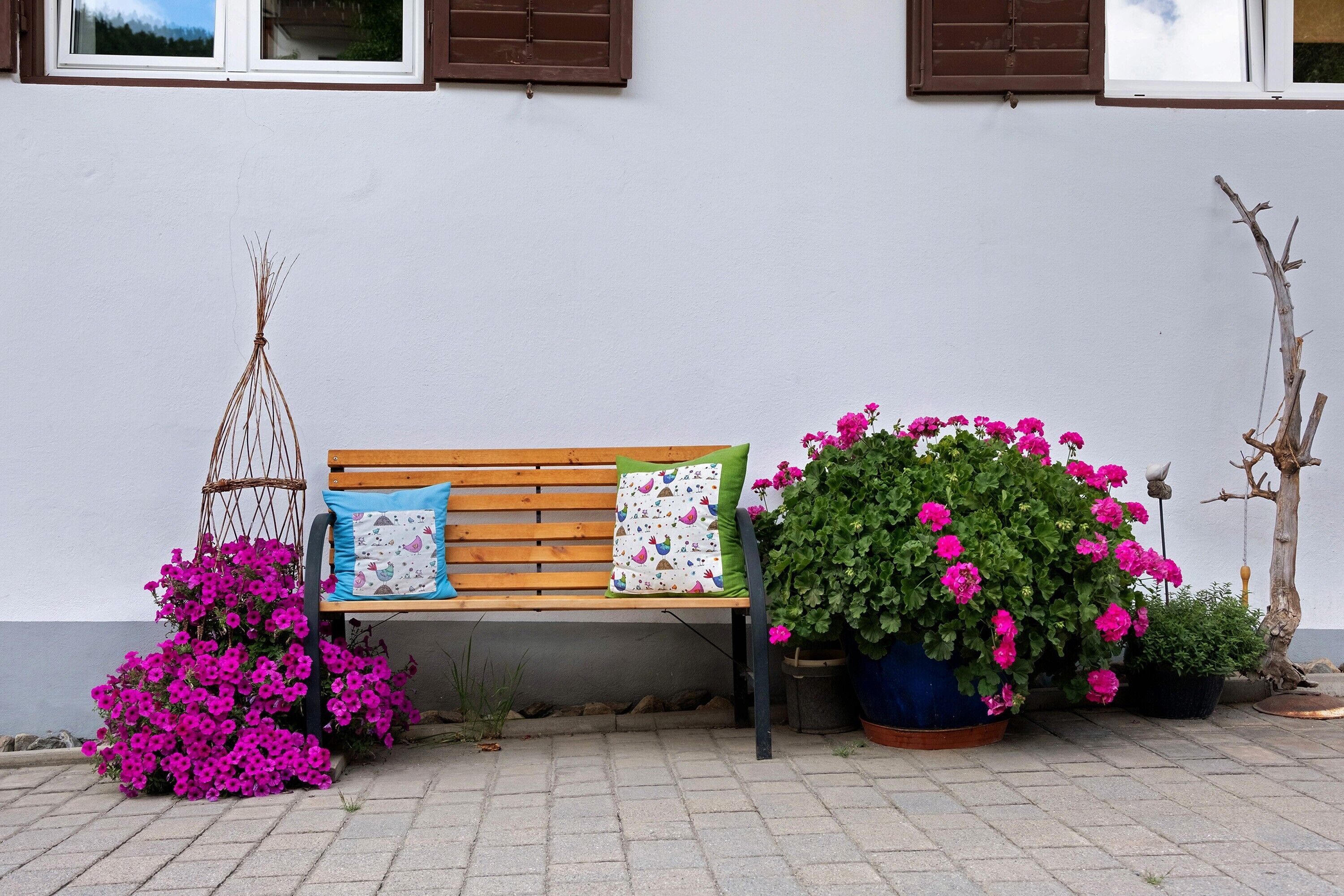 A bench with two cushions, a hanging basket with pink flowers, and a large potted plant with pink flowers.