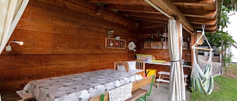 A wooden cabin with a bed, a table, and a bench. There is a window with curtains, a hanging chair, and a small shelf with items.