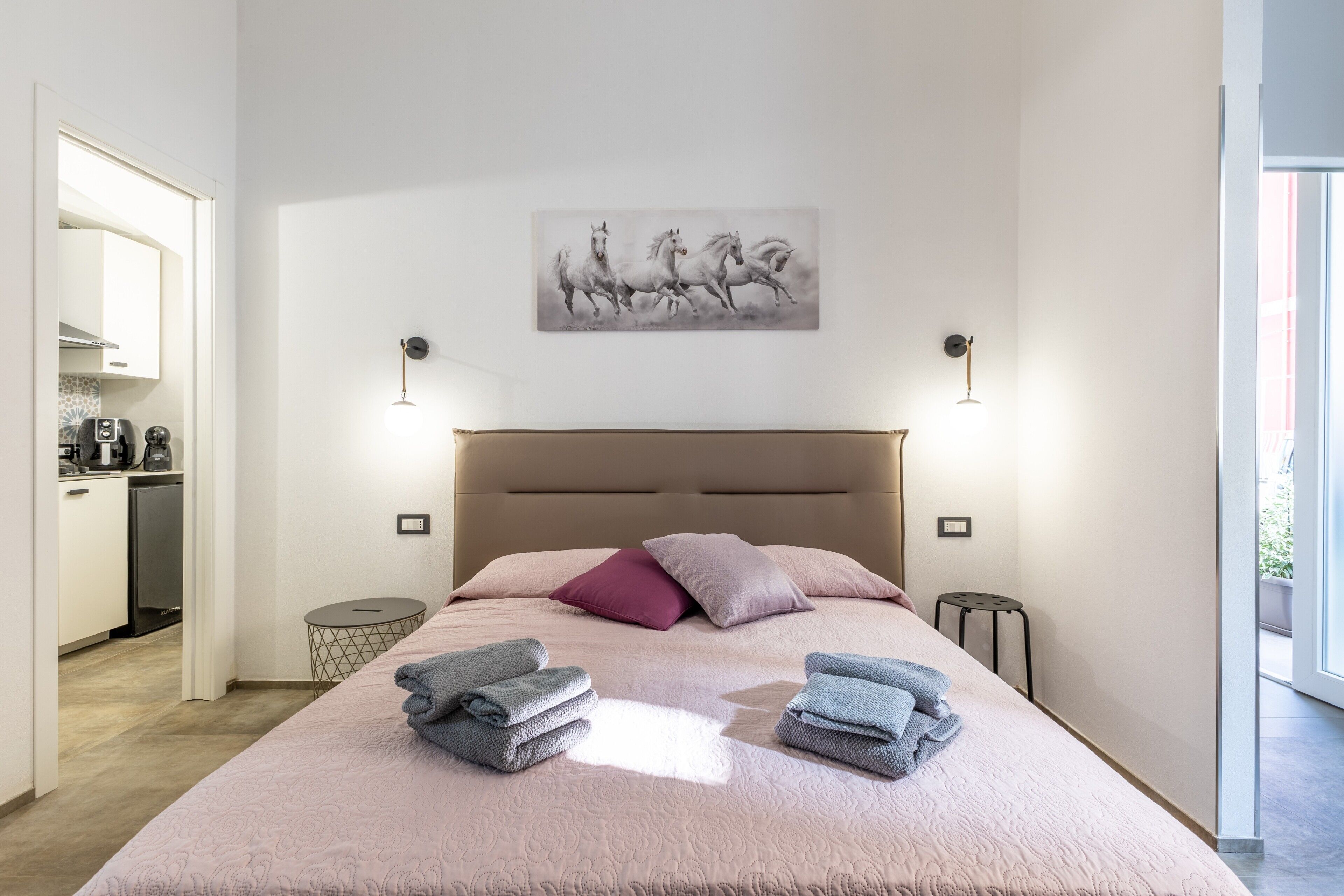 A bedroom with a bed, pillows, a painting, bedside lamps, and a view of the kitchen.