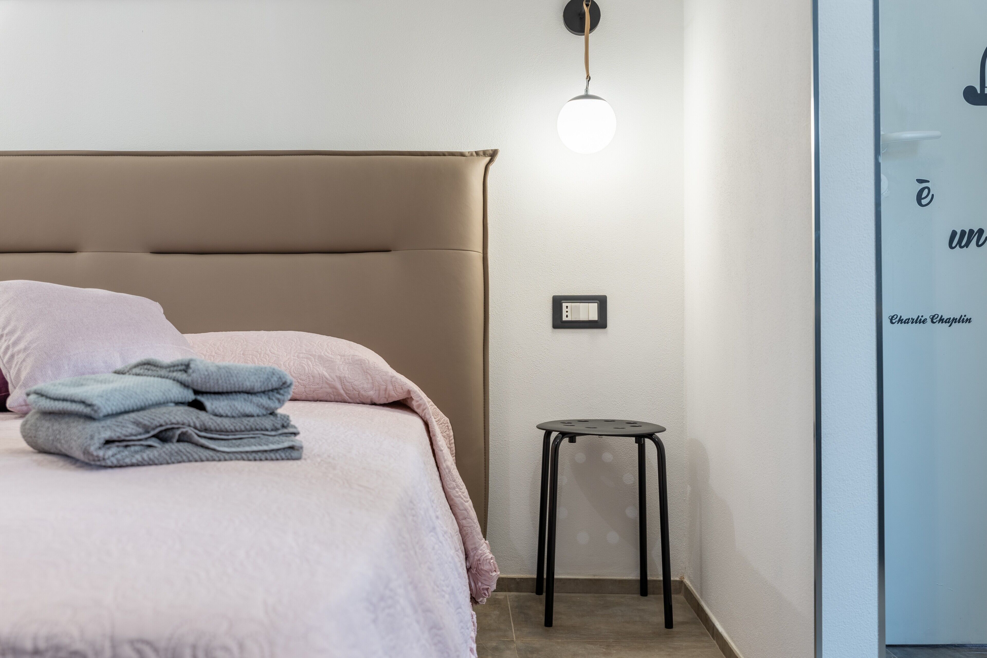 Un dormitorio con cama, un taburete pequeño, un enchufe de pared y una lámpara.