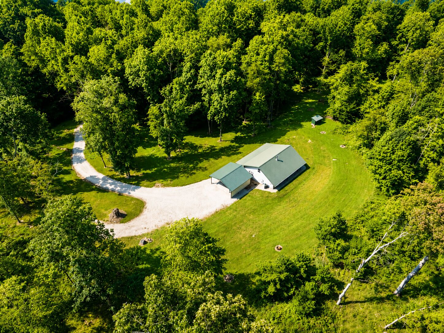 A large, single-story building with a green roof, surrounded by dense forest. A gravel driveway leads to the entrance.