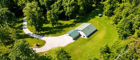 A large, single-story building with a green roof, surrounded by dense forest. A gravel driveway leads to the entrance.