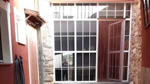 Un'area patio esterna con pavimento in pietra, due panche in legno, un'ampia finestra con tende e un muro in pietra.