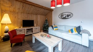 Un salón con techo de madera, un televisor de pantalla plana, un sofá blanco, una silla roja, una mesa de centro y un árbol de Navidad.