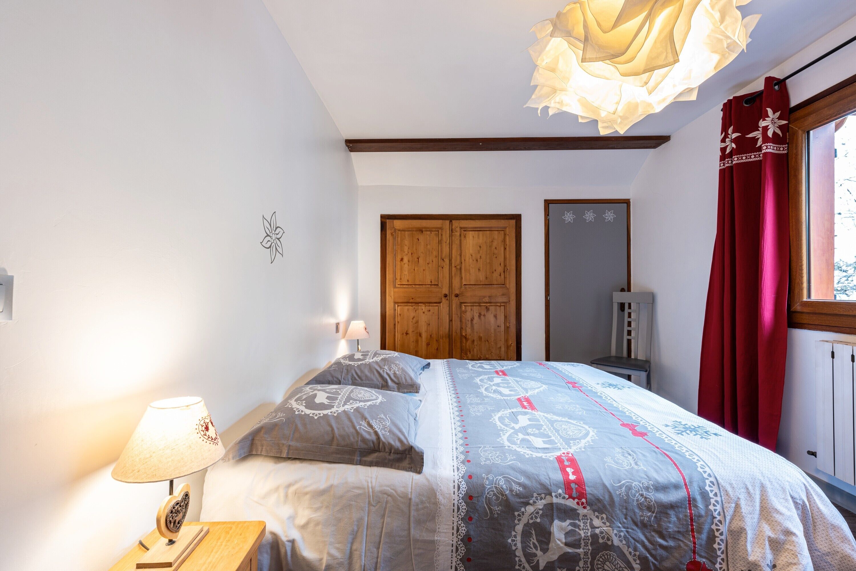 Un dormitorio con cama, armario de madera, mesita de noche con lámpara y ventana con cortinas.