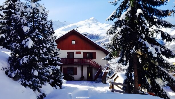 Un albergue en la montaña cubierto de nieve, con tejado rojo, rodeado de árboles cargados de nieve y un cielo despejado.