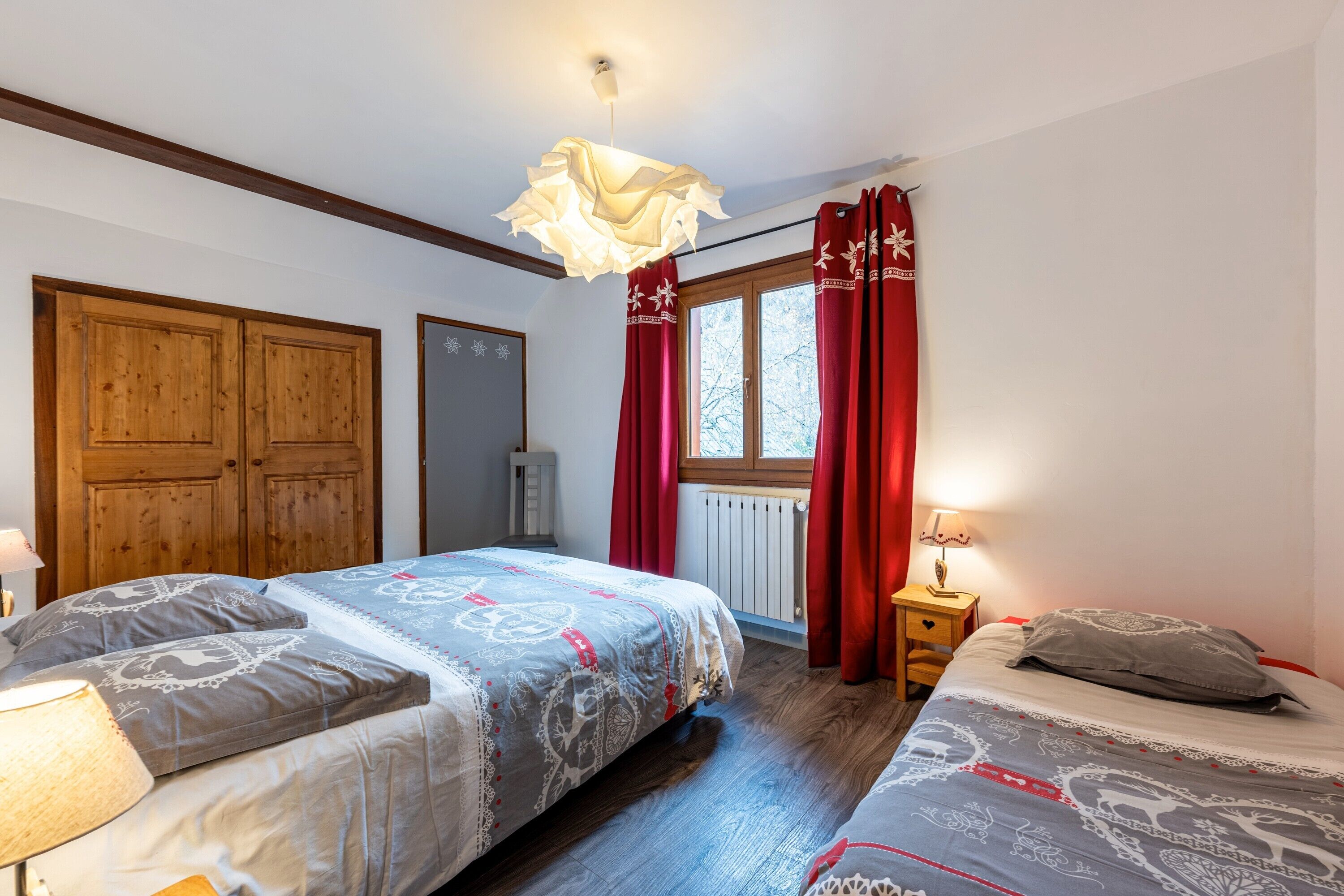 A bedroom with two beds, wooden wardrobe, window with red curtains, and a hanging light fixture.
