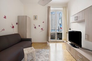 Un salon comprenant un canapé, une petite table, un téléviseur posé sur un meuble et une porte donnant sur un balcon.