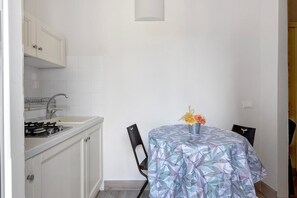 Una pequeña cocina con gabinetes blancos, una estufa a gas y un lavamanos. Hay una mesa de comedor con mantel floreado y dos sillas.