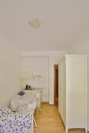 Une petite pièce lumineuse, avec un lit, une table entourée de chaises, une armoire et une porte donnant sur une autre pièce.