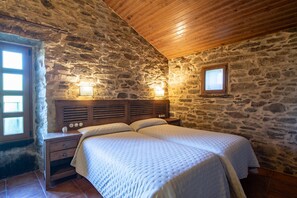 Une chambre avec un mur en pierre, un plafond en bois, un lit avec une tête de lit, des lampes de chevet et une fenêtre.
