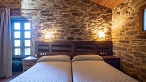 Ein Zimmer mit Steinwand, Holzboden, zwei Betten mit Kopfteilen, einem Nachttisch, einem Fenster mit Vorhängen und Wandleuchten.