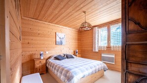 Un dormitorio de cabaña de madera con una cama, mesitas de noche, un banco, un ventilador de techo y una ventana con cortinas.
