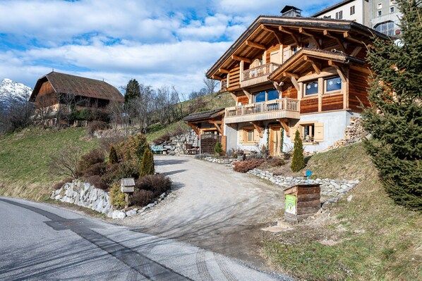 A wooden chalet with a balcony, a stone wall, and a mountain backdrop.