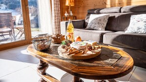 Una mesa de madera con una bandeja de quesos, una botella y una tetera en una habitación con un sofá y vistas a las montañas.