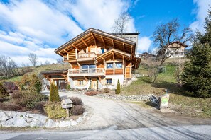 A large wooden chalet with a balcony, a stone pathway, and a mountain backdrop.