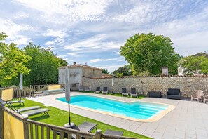 A swimming pool with lounge chairs, a stone wall, and a small building with a tiled roof.