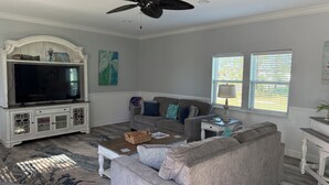 Una sala de estar con un sofá gris, una mesa de centro, un televisor sobre un mueble blanco, un ventilador de techo y dos ventanas con persianas.