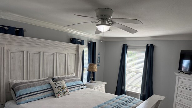Un dormitorio con cama, ventilador de techo, mesita de noche, cómoda y ventana con cortinas.