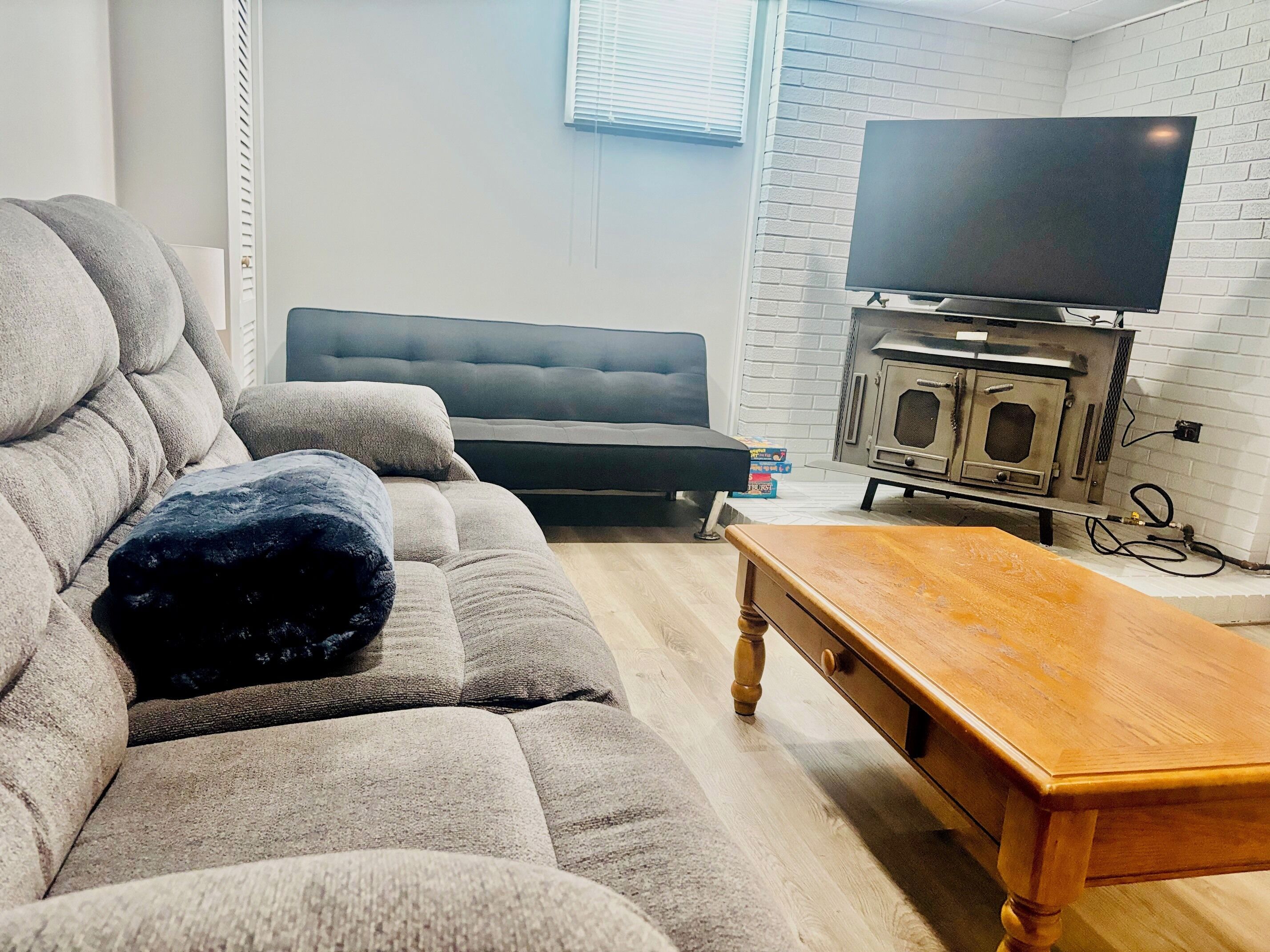 Een woonkamer met een grijze bank, een houten salontafel, een zwart voetenbankje, een zwarte fauteuil en een televisie op een dressoir.