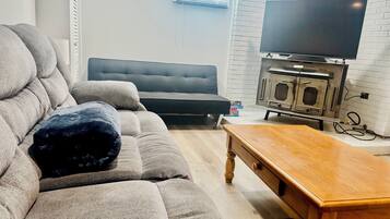 Een woonkamer met een grijze bank, een houten salontafel, een zwart voetenbankje, een zwarte fauteuil en een televisie op een dressoir.
