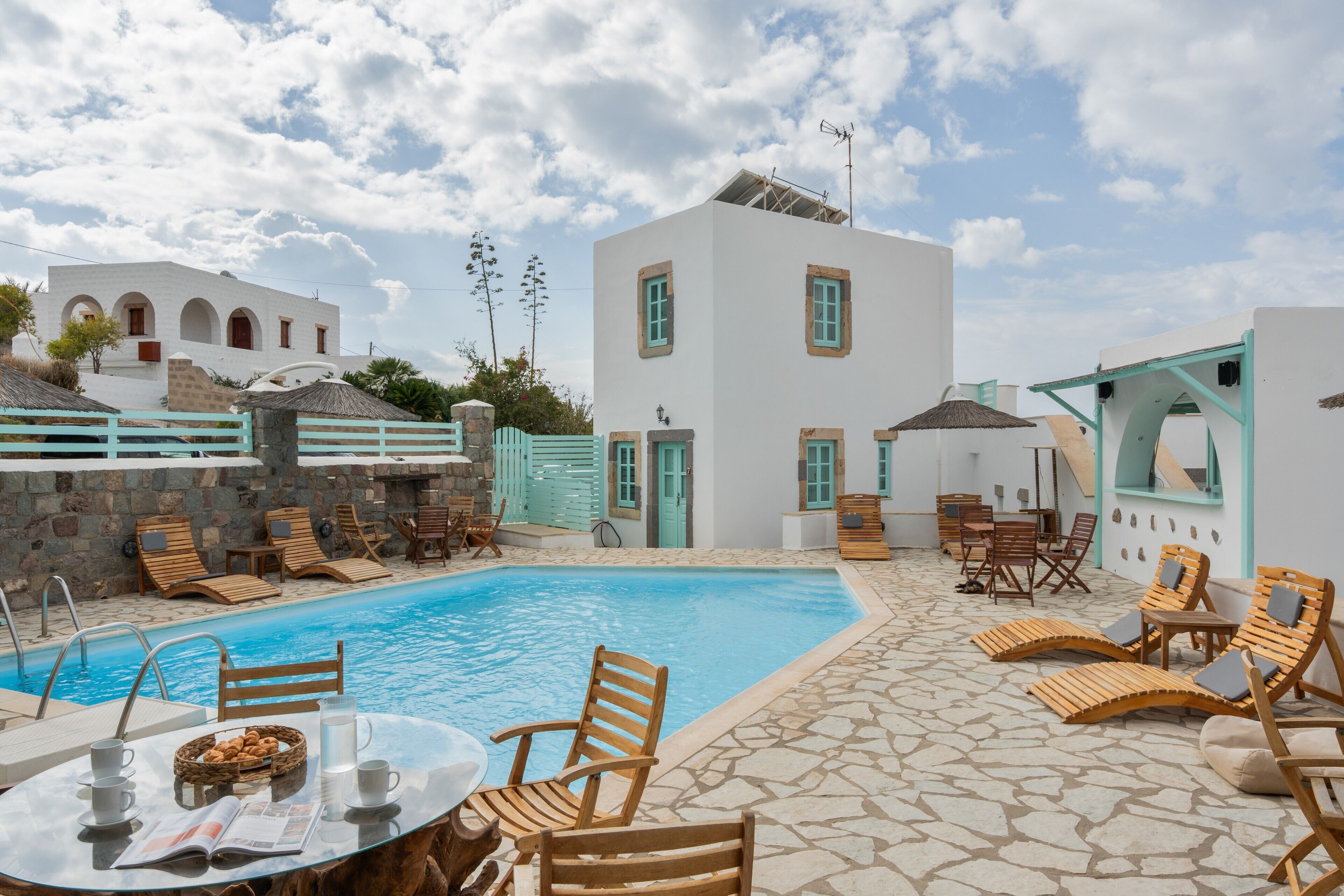 Un espace aménagé au bord de la piscine, avec des chaises longues, une table où sont servis des boissons et un bâtiment doté de fenêtres en arc de cercle.