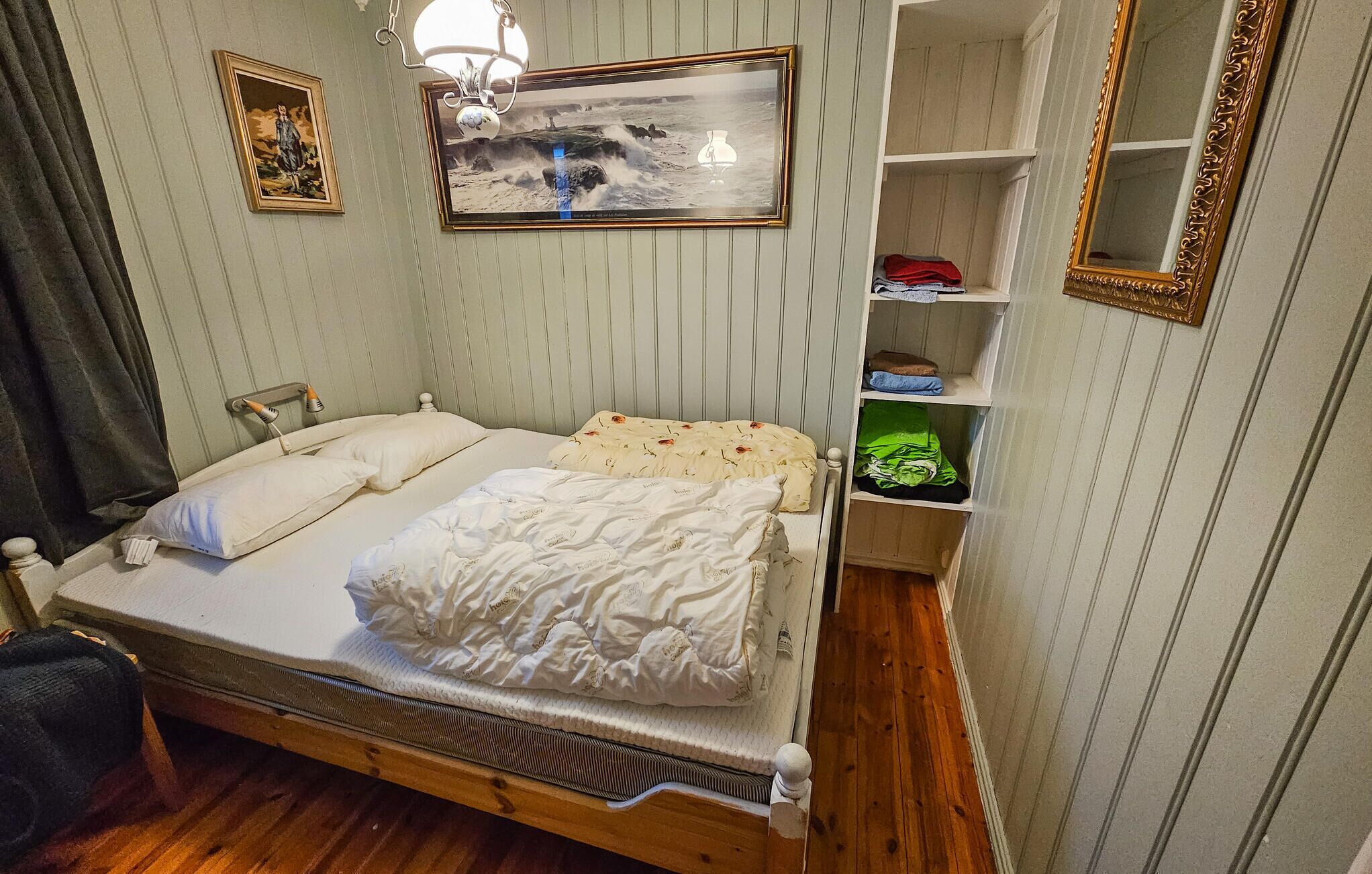 A neatly made bed with a wooden frame, a bedside table with a lamp, a shelf with folded clothes, and a framed picture on the wall.