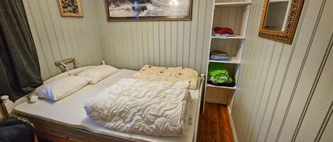 A neatly made bed with a wooden frame, a bedside table with a lamp, a shelf with folded clothes, and a framed picture on the wall.