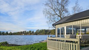 Eine Blockhütte am Seeufer mit einer Terrasse mit Blick auf das Wasser, großen Fenstern und Blick auf den umliegenden Wald.