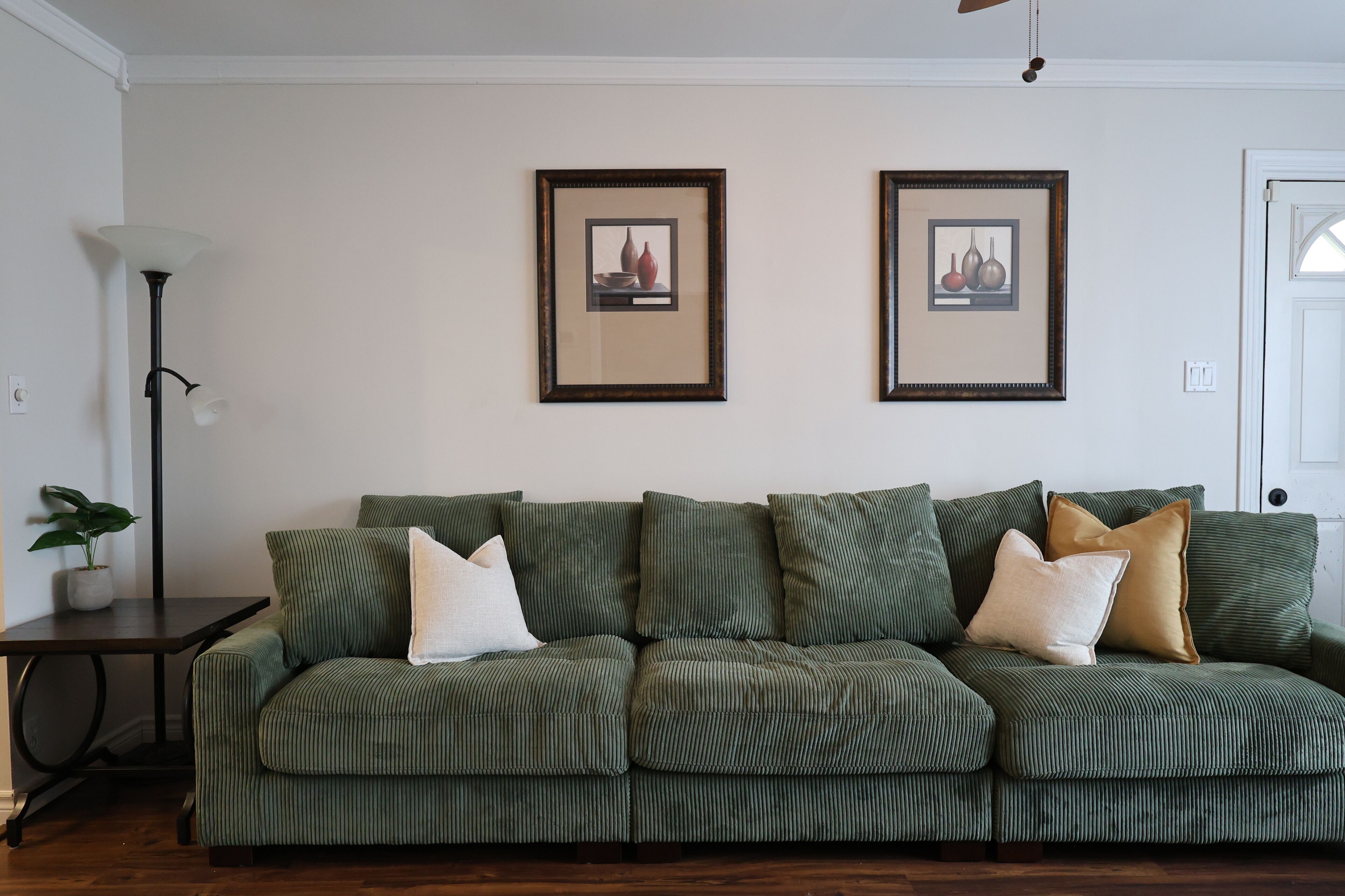 A green upholstered sofa with multiple cushions, a dark wooden side table with a potted plant, and two framed artworks on the wall.