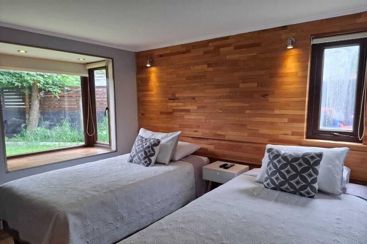 Ein Schlafzimmer mit zwei Betten, einem Holzkopfteil, einem Nachttisch und einem Fenster mit Blick auf Grünflächen.
