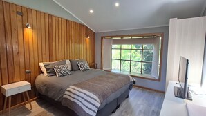 Un dormitorio con una cama grande, paneles de madera en las paredes, una ventana con vistas a la vegetación y un escritorio con una computadora.
