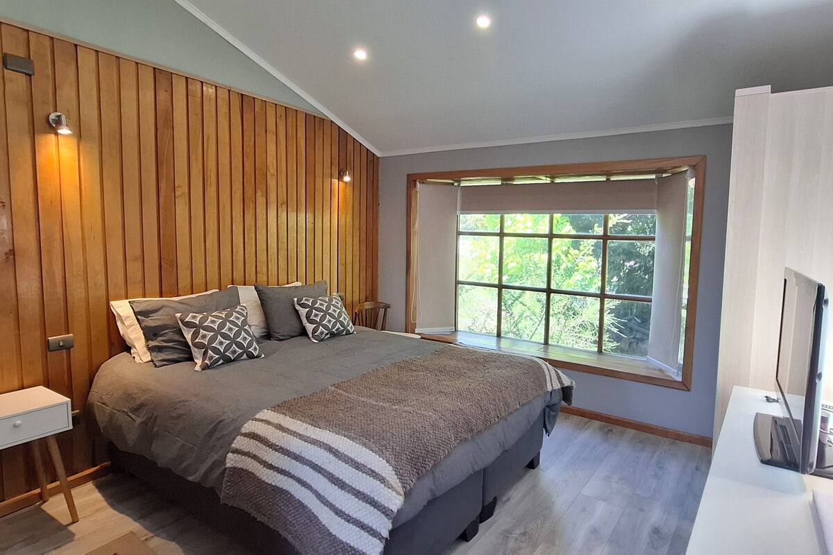 Ein Schlafzimmer mit einem großen Bett, Holzvertäfelung an den Wänden, einem Fenster mit Blick ins Grüne und einem Schreibtisch mit Computer.